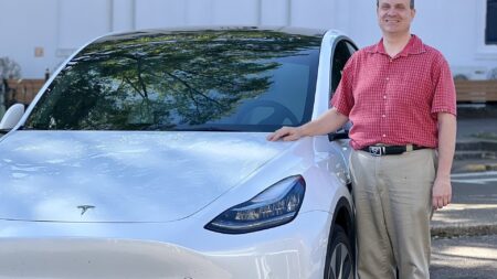 Bill Taylor, dressed in a red shirt and khaki pants, poses alongside his white Tesla Model Y on the Square.